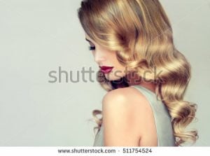 Profile view of a woman with long, wavy blonde hair and red lipstick against a neutral background.