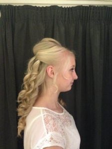 Side profile of a woman with long, curly blonde hair styled elegantly against a dark curtain.