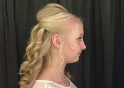 Side profile of a woman with long, curly blonde hair styled elegantly against a dark curtain.