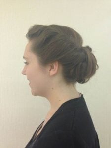 Side profile of a woman with brown hair styled in a low bun updo, wearing a black top against a neutral background, typical for a salon gallery