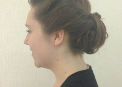 Side profile of a woman with brown hair styled in a low bun updo, wearing a black top against a neutral background, typical for a salon gallery