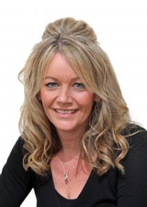 Woman with long, curly blonde hair styled in a half-updo, wearing a black top and smiling at the camera.