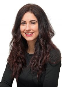 Woman with long, curly dark hair wearing a black top, smiling at the camera against a white background.