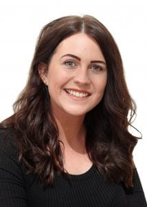 Woman with long brown hair and a black top smiling at the camera in a neutral setting.