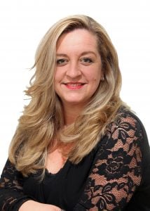 Woman with long blonde hair styled in loose waves, wearing a black lace top and smiling at the camera.