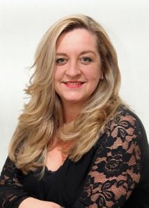 A woman with long blonde hair wearing a black top with lace details, smiling at the camera.