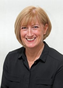 Woman with short blonde hair wearing a black shirt, smiling at the camera with a neutral background.